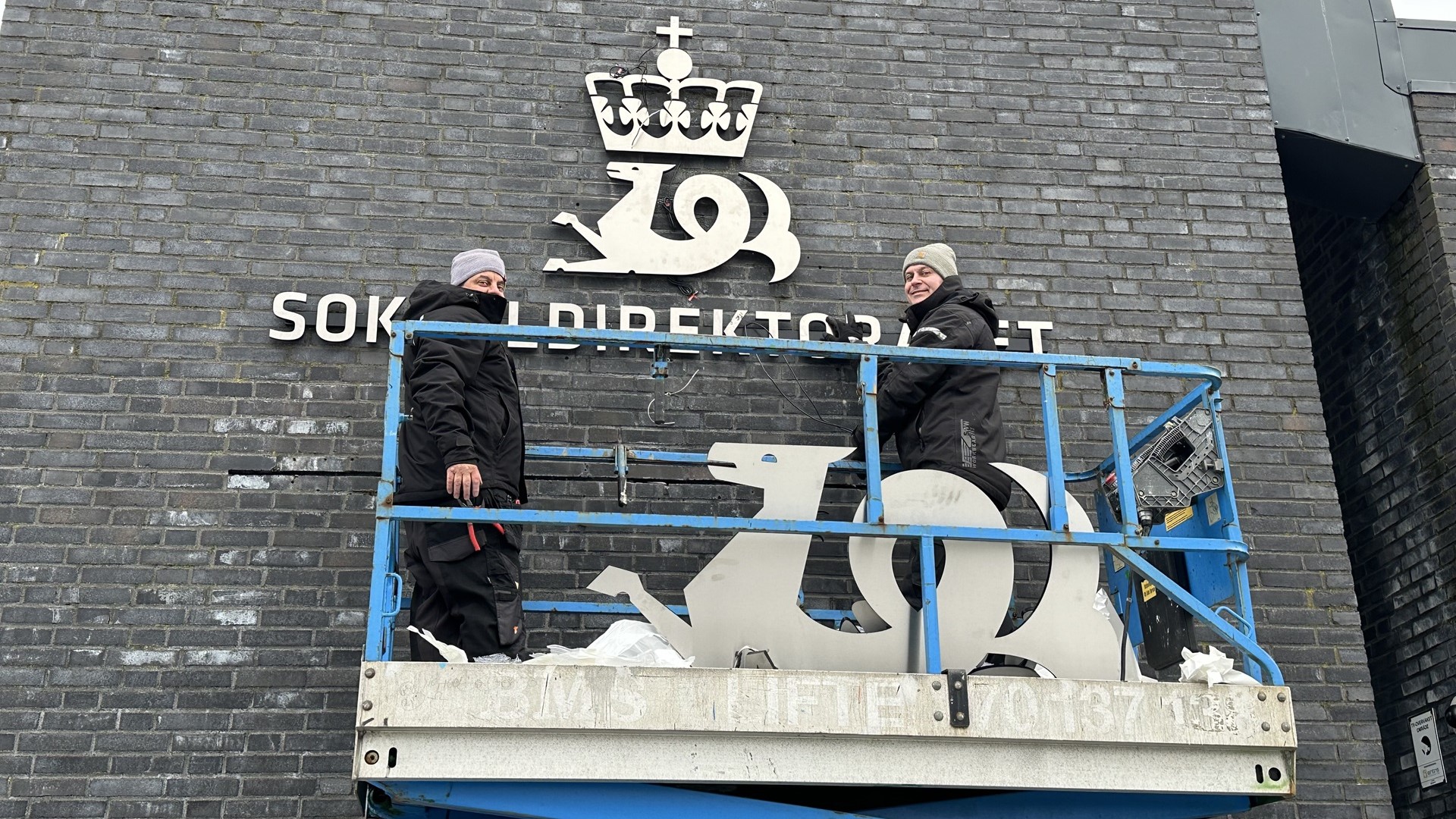 To menn står i en lift for å henge opp et skilt.